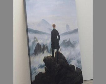 May include: A rectangular canvas print featuring a painting of a person in a dark coat standing on a rocky cliff edge. The person holds a walking stick and looks out over a vast, misty mountain landscape with jagged peaks emerging from clouds.