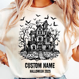 A woman wearing a white t-shirt with a black and white illustration of a spooky house surrounded by bats and pumpkins, along with the text "CUSTOM NAME HALLOWEEN 2025".