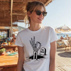 May include: White t-shirt featuring a black graphic of a skeleton hand making the "okay" gesture, emerging from a cracked surface. A tombstone with "I'M OKAY" is next to the hand. The shirt is a casual, everyday wear item.