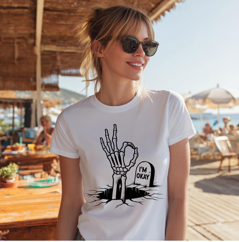 May include: White t-shirt featuring a black graphic of a skeleton hand making the "okay" gesture, emerging from a cracked surface. A tombstone with "I'M OKAY" is next to the hand. The shirt is a casual, everyday wear item.
