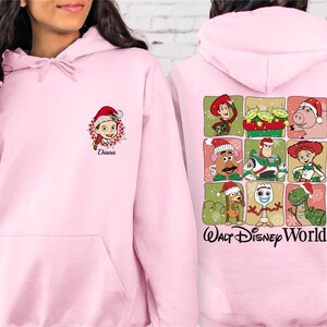 Puede incluir: Sudadera con capucha rosa claro. La parte delantera presenta un personaje de dibujos animados con un gorro de Pap&aacute; Noel y el nombre "Diana". La parte trasera muestra una cuadr&iacute;cula de personajes de Toy Story con gorros de Pap&aacute; Noel, con "Walt Disney World".