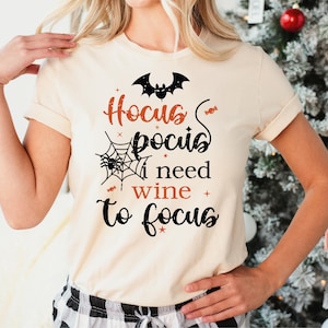 May include: A cream-colored short-sleeve t-shirt featuring a black and orange graphic print that reads Hocus Pocus I need wine to focus. The design includes a black bat, a spider web with a spider, and small orange candy icons scattered around the text.