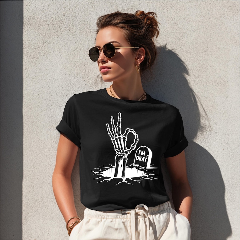 May include: Black t-shirt with a white graphic of a skeleton hand making the "okay" gesture emerging from a crack in the ground. A tombstone with "I'M OKAY" is also in the graphic. The shirt is a casual wear item.