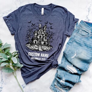 A dark blue t-shirt with a Halloween-themed graphic and the text "CUSTOM NAME HALLOWEEN 2015" printed on it, along with a pair of distressed blue jeans and some greenery.