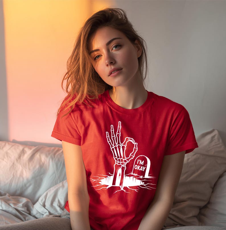 May include: Red t-shirt featuring a white graphic of a skeleton hand making the "okay" gesture, with a tombstone that reads "I'M OKAY". The shirt is short-sleeved and has a crew neck.
