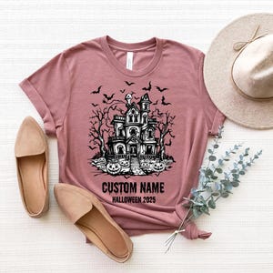 a pink t-shirt with a Halloween-themed graphic, a beige hat, a pair of beige shoes, and some greenery, all laid out on a white surface.