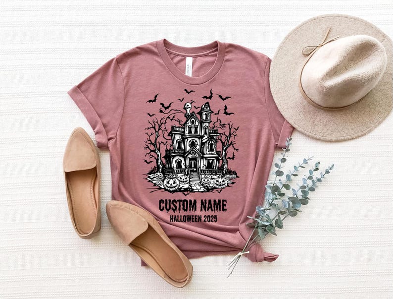 a pink t-shirt with a Halloween-themed graphic, a beige hat, a pair of beige shoes, and some greenery, all laid out on a white surface.