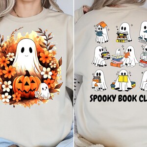 Peut inclure: Sweat-shirt beige avec un motif sur le thème d'Halloween. L'avant présente un fantôme souriant, une citrouille et des fleurs. Le dos affiche des illustrations de fantômes lisant des livres, avec le texte "Spooky Book Club" en noir.