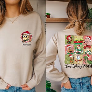 Puede incluir: Sudadera beige con un dibujo de Slinky Dog con un gorro de Pap&aacute; Noel y el nombre "Adrienne". La parte trasera tiene una cuadr&iacute;cula de personajes de Toy Story con gorros de Pap&aacute; Noel, con el texto "Walt Disney World".