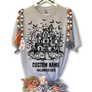 A grey t-shirt with a Halloween-themed graphic design, including a house, bats, and pumpkins. The t-shirt is hanging on a hanger, and there are Halloween-themed beads and decorations around it.