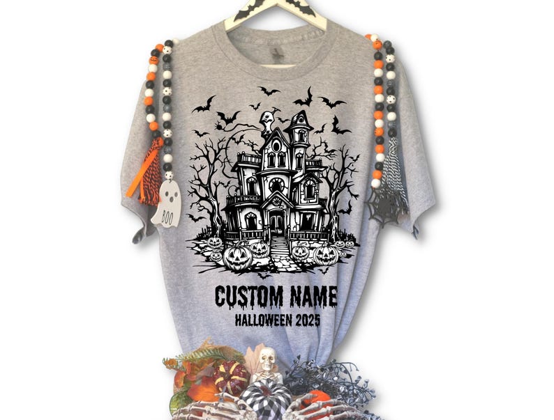 A grey t-shirt with a Halloween-themed graphic design, including a house, bats, and pumpkins. The t-shirt is hanging on a hanger, and there are Halloween-themed beads and decorations around it.