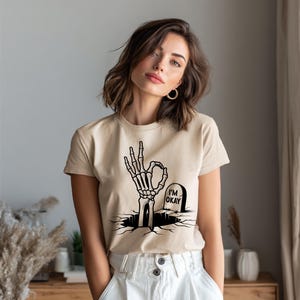 May include: Beige t-shirt featuring a black graphic of a skeleton hand making the "okay" gesture, emerging from a cracked ground. A tombstone with "I'M OKAY" is next to the hand. The shirt is paired with white pants.