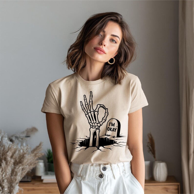 May include: Beige t-shirt featuring a black graphic of a skeleton hand making the "okay" gesture, emerging from a cracked ground. A tombstone with "I'M OKAY" is next to the hand. The shirt is paired with white pants.
