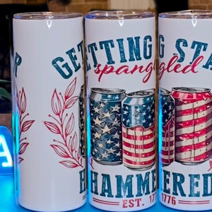 May include: White stainless steel tumblers with patriotic designs. One tumbler has the text "Getting Star Spangled Hammered" with images of beer cans in red, white, and blue. Another has the text "Sip" with red floral accents.