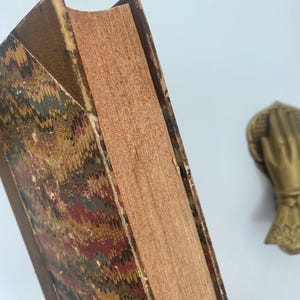 Puede incluir: Primer plano de un libro antiguo con una cubierta jaspeada en tonos rojos, dorados y verdes. Las p&aacute;ginas envejecidas tienen una textura rugosa y el lomo est&aacute; desgastado. Una mano decorativa de lat&oacute;n es visible en el fondo.