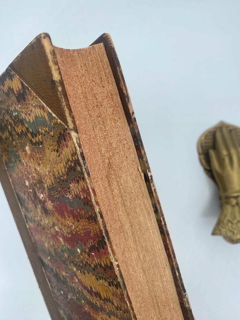 Puede incluir: Primer plano de un libro antiguo con una cubierta jaspeada en tonos rojos, dorados y verdes. Las p&aacute;ginas envejecidas tienen una textura rugosa y el lomo est&aacute; desgastado. Una mano decorativa de lat&oacute;n es visible en el fondo.