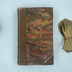 Puede incluir: Un libro antiguo con una cubierta jaspeada en tonos rojos, marrones, amarillos y verdes. El lomo es de color marr&oacute;n liso y los bordes muestran signos de desgaste. Se ve un llamador de puerta decorativo de lat&oacute;n con forma de mano.