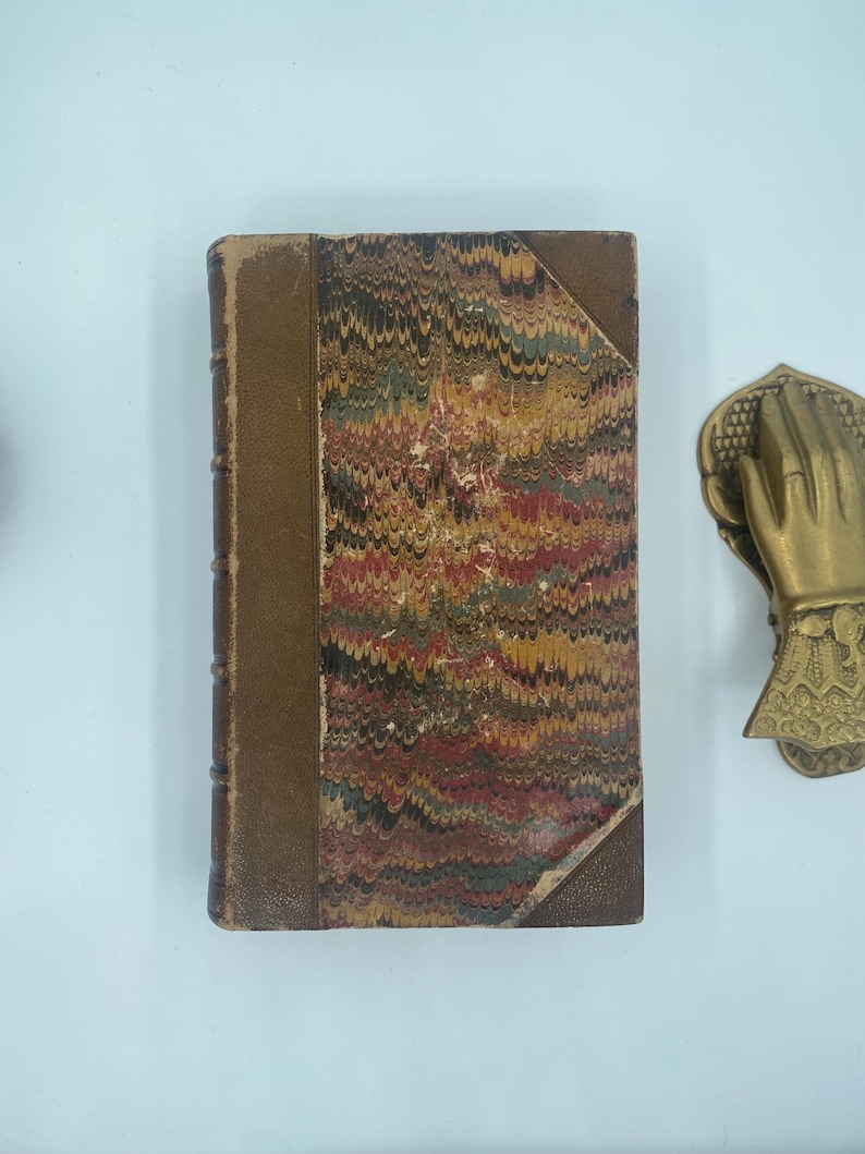 Puede incluir: Un libro antiguo con una cubierta jaspeada en tonos rojos, marrones, amarillos y verdes. El lomo es de color marr&oacute;n liso y los bordes muestran signos de desgaste. Se ve un llamador de puerta decorativo de lat&oacute;n con forma de mano.