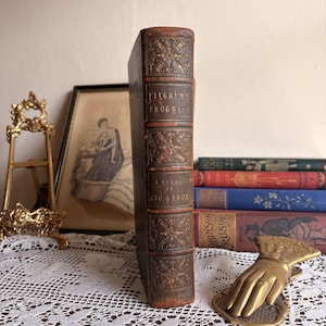 May include: A vintage leather-bound book titled Pilgrim's Progress by Geo. Offor with gold-embossed floral spine details, standing upright next to a stack of three colorful books and a brass hand-shaped paperweight on a white lace cloth.