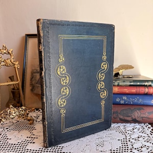 Peut inclure: Un livre à couverture rigide bleu foncé avec des motifs en filigrane dorés en relief sur la couverture et le dos. Le livre est placé à la verticale, présentant un motif de bordure rectangulaire avec des volutes décoratives sur les côtés.