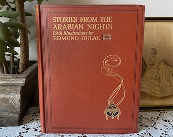 May include: A vintage hardcover book titled "Stories From The Arabian Nights" by Edmund Dulac. The book has a reddish-brown cover with gold lettering and an ornate illustration. The book is displayed on a white lace tablecloth.