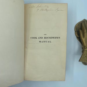 Puede incluir: Libro abierto con el t&iacute;tulo "THE COOK AND HOUSEWIFE'S MANUAL" impreso en la p&aacute;gina derecha. Texto manuscrito visible en la parte superior de la p&aacute;gina. Un objeto decorativo de lat&oacute;n en forma de mano est&aacute; en el lado derecho de la imagen.