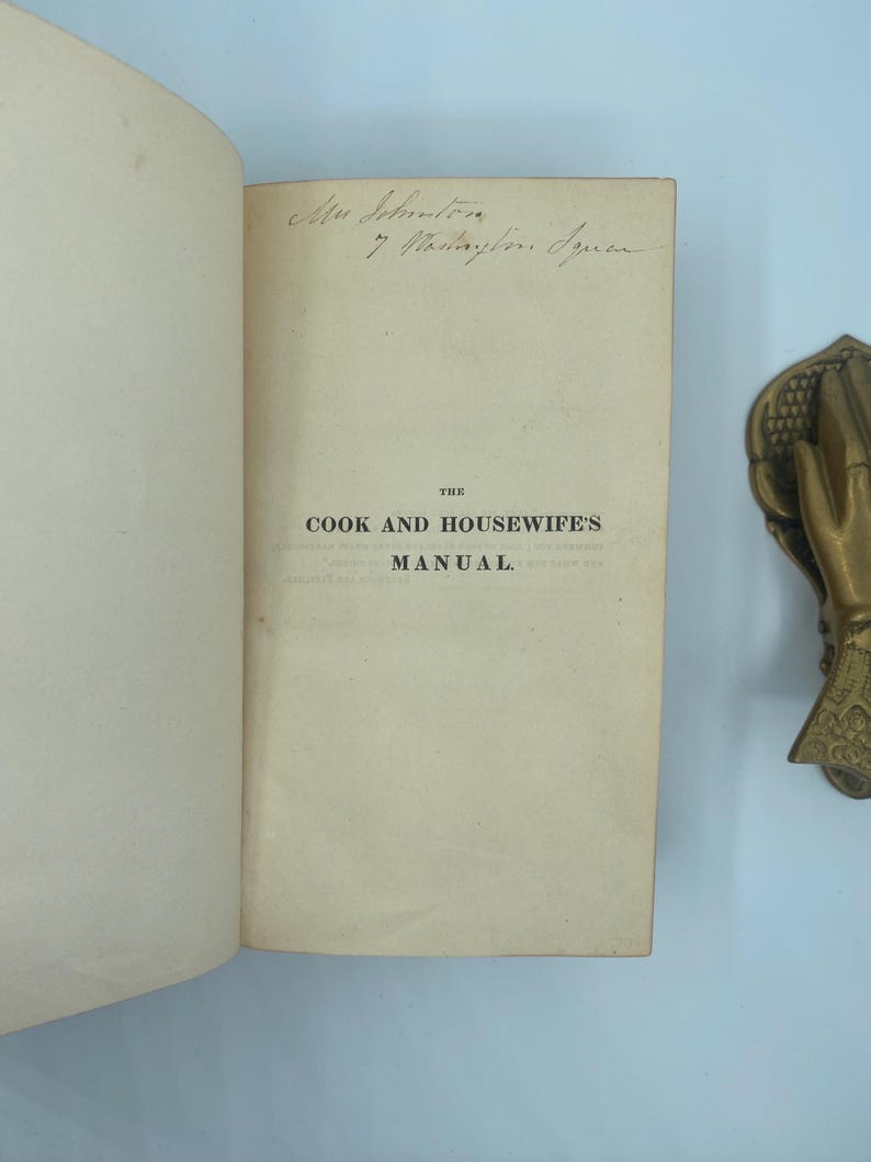 Puede incluir: Libro abierto con el t&iacute;tulo "THE COOK AND HOUSEWIFE'S MANUAL" impreso en la p&aacute;gina derecha. Texto manuscrito visible en la parte superior de la p&aacute;gina. Un objeto decorativo de lat&oacute;n en forma de mano est&aacute; en el lado derecho de la imagen.