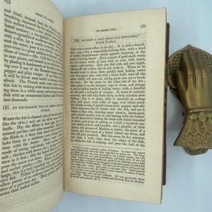 Puede incluir: Libro abierto con recetas de pescado, con un sujetalibros en forma de mano de lat&oacute;n. El libro est&aacute; abierto en las p&aacute;ginas 172 y 173, con texto sobre fondo blanco. El libro est&aacute; en buen estado.