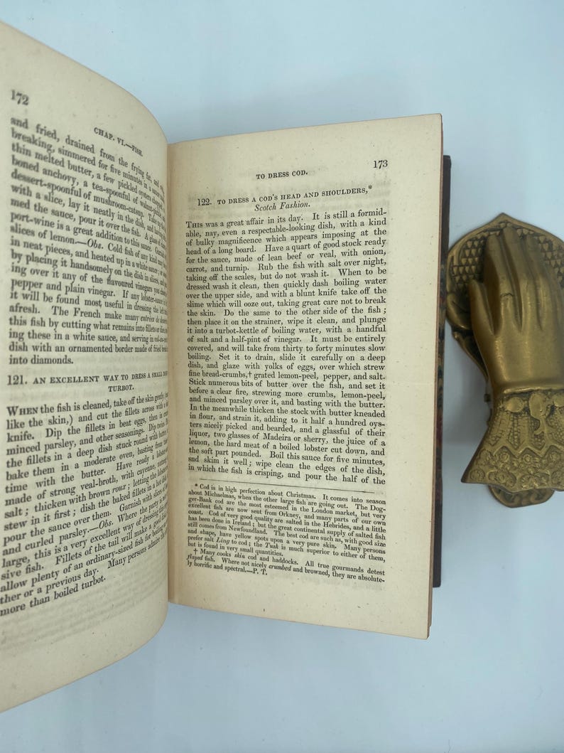 Puede incluir: Libro abierto con recetas de pescado, con un sujetalibros en forma de mano de lat&oacute;n. El libro est&aacute; abierto en las p&aacute;ginas 172 y 173, con texto sobre fondo blanco. El libro est&aacute; en buen estado.