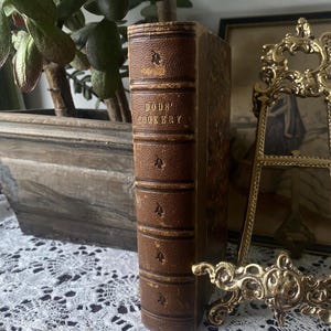 Puede incluir: Un libro de cocina antiguo titulado "BODS' COOKERY" con una cubierta de cuero marr&oacute;n. La columna vertebral est&aacute; grabada con el t&iacute;tulo y elementos decorativos. El libro est&aacute; de pie sobre una superficie de encaje blanco, con una planta y un marco decorativo en el fondo.