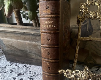 Op de afbeelding: Een vintage kookboek getiteld "BODS' COOKERY" met een bruine leren kaft. De rug is in reliëf met de titel en decoratieve elementen. Het boek staat rechtop op een wit kanten oppervlak, met een plant en een decoratief frame op de achtergrond.