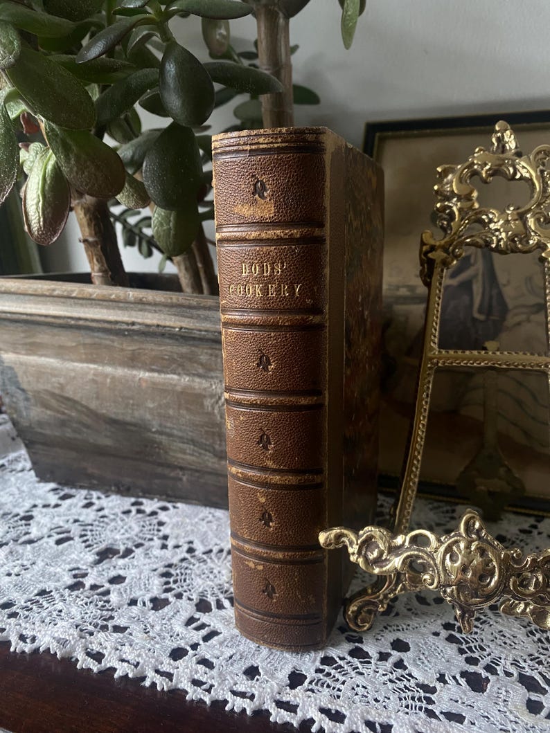 Puede incluir: Un libro de cocina antiguo titulado "BODS' COOKERY" con una cubierta de cuero marr&oacute;n. La columna vertebral est&aacute; grabada con el t&iacute;tulo y elementos decorativos. El libro est&aacute; de pie sobre una superficie de encaje blanco, con una planta y un marco decorativo en el fondo.