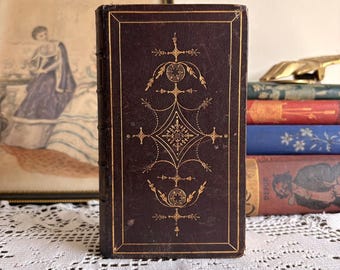 May include: A dark brown hardcover book featuring intricate gold-colored filigree and geometric patterns on the front cover. The spine has raised bands, and the book is positioned upright in front of a stack of other books and a framed illustration.