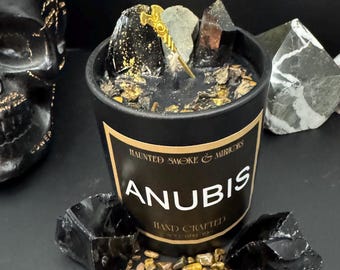May include: Black candle with gold lettering that reads "Haunted Smoke & Mirrors Anubis Hand Crafted". The candle is surrounded by black and gold stones.