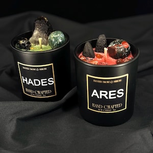 May include: Two black soy candles with gold labels, one labeled "HADES" and the other "ARES". The Hades candle has a green wax base with crystals and a black stone. The Ares candle has a red wax base with crystals and black incense cones.