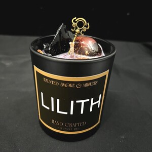 May include: Black candle jar with a gold label that reads "Haunted Smoke & Mirrors Lilith Hand Crafted Soy Candle 10oz". The candle has a black and red crystal and a gold key charm on top.