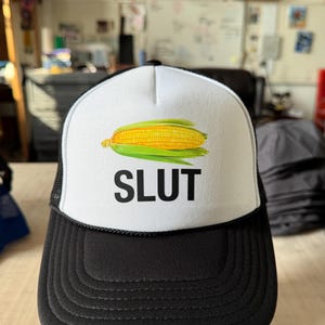 May include: A black and white trucker hat with a white front panel. The front panel features a yellow corn graphic and the word "SLUT" in black block letters. The hat has a mesh back.