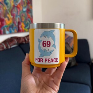 May include: Yellow insulated mug with a silver rim and a handle. The mug features a sticker with two blue dolphins, the number 69, and the words "FOR PEACE".