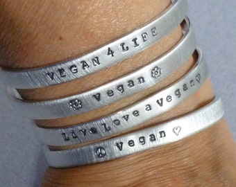May include: Five silver bracelets with vegan-themed messages. The bracelets are stacked on a person's wrist. The messages on the bracelets are "Vegan 4 Life", "Vegan", "Live Love a Vegan", and "Vegan".