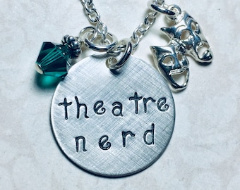 May include: Silver necklace with a round charm that says "theatre nerd" and two silver mask charms. The necklace also has a green crystal charm.
