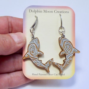May include: A pair of laser cut wood shark earrings with white painted details. The earrings are on a card that says "Dolphin Moon Creations" and "Hand Painted Laser Cut Wood".