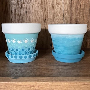 May include: Two terracotta plant pots painted in a blue ombre design. One pot has a white polka dot pattern and the other has a white brushstroke pattern.