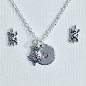 May include: Silver necklace with a silver turtle charm and a silver disc charm with the letter 'D' engraved on it. The necklace comes with two smaller silver turtle charms.