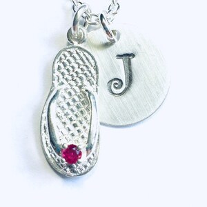 May include: Silver necklace with a flip-flop charm and a round charm with the letter J. The flip-flop charm has a small red gemstone.