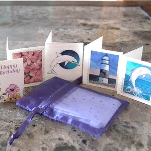 May include: Five small cards with different images on them. The cards are arranged in a row, with a purple satin bag in front of them. The cards feature images of a dolphin, a lighthouse, a flower, and a happy birthday message.