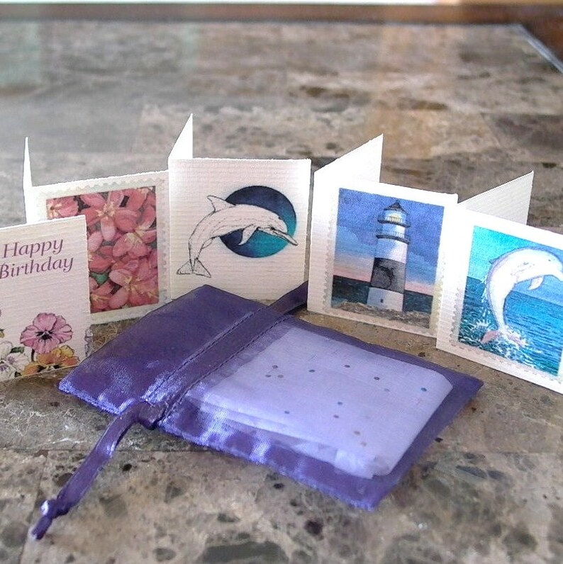 May include: Five small cards with different images on them. The cards are arranged in a row, with a purple satin bag in front of them. The cards feature images of a dolphin, a lighthouse, a flower, and a happy birthday message.