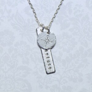 May include: Silver necklace with a compass charm and a bar pendant with the word "erlust" engraved on it.