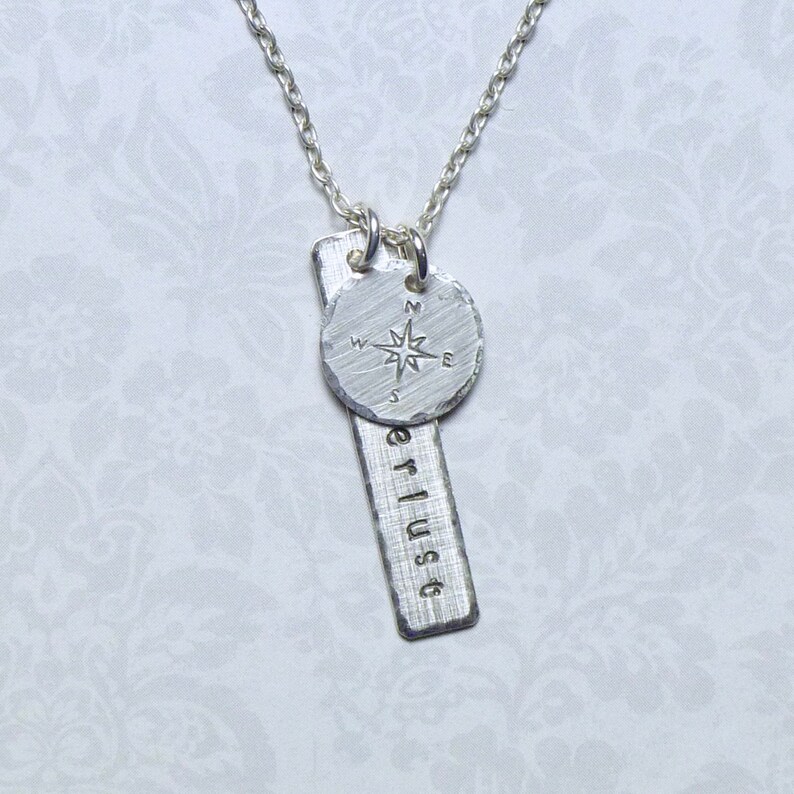 May include: Silver necklace with a compass charm and a bar pendant with the word "erlust" engraved on it.
