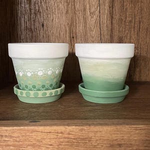 May include: Two white ceramic plant pots with green and white painted designs. The pots have a green and white ombre effect and are sitting on a wooden surface.