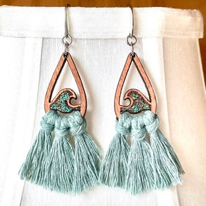 May include: Teal blue tassel earrings with a wooden teardrop shaped pendant featuring a glitter wave design.